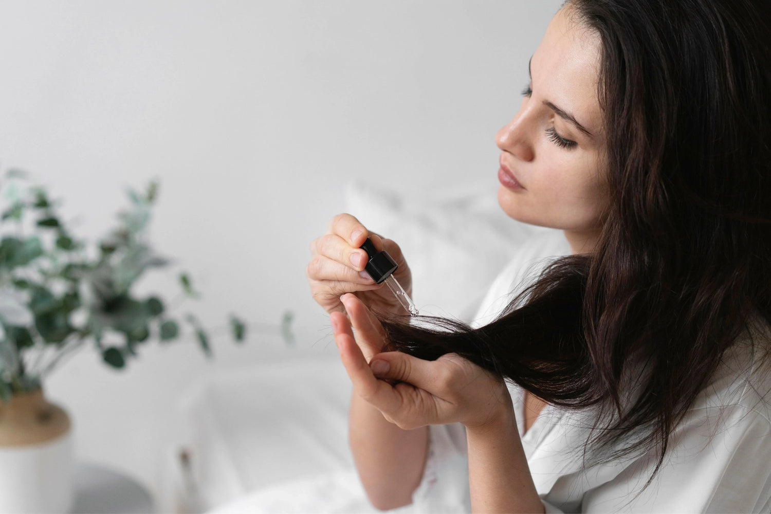 Woman applying hair serum with dropper on dark hair in bright, minimalist indoor setting