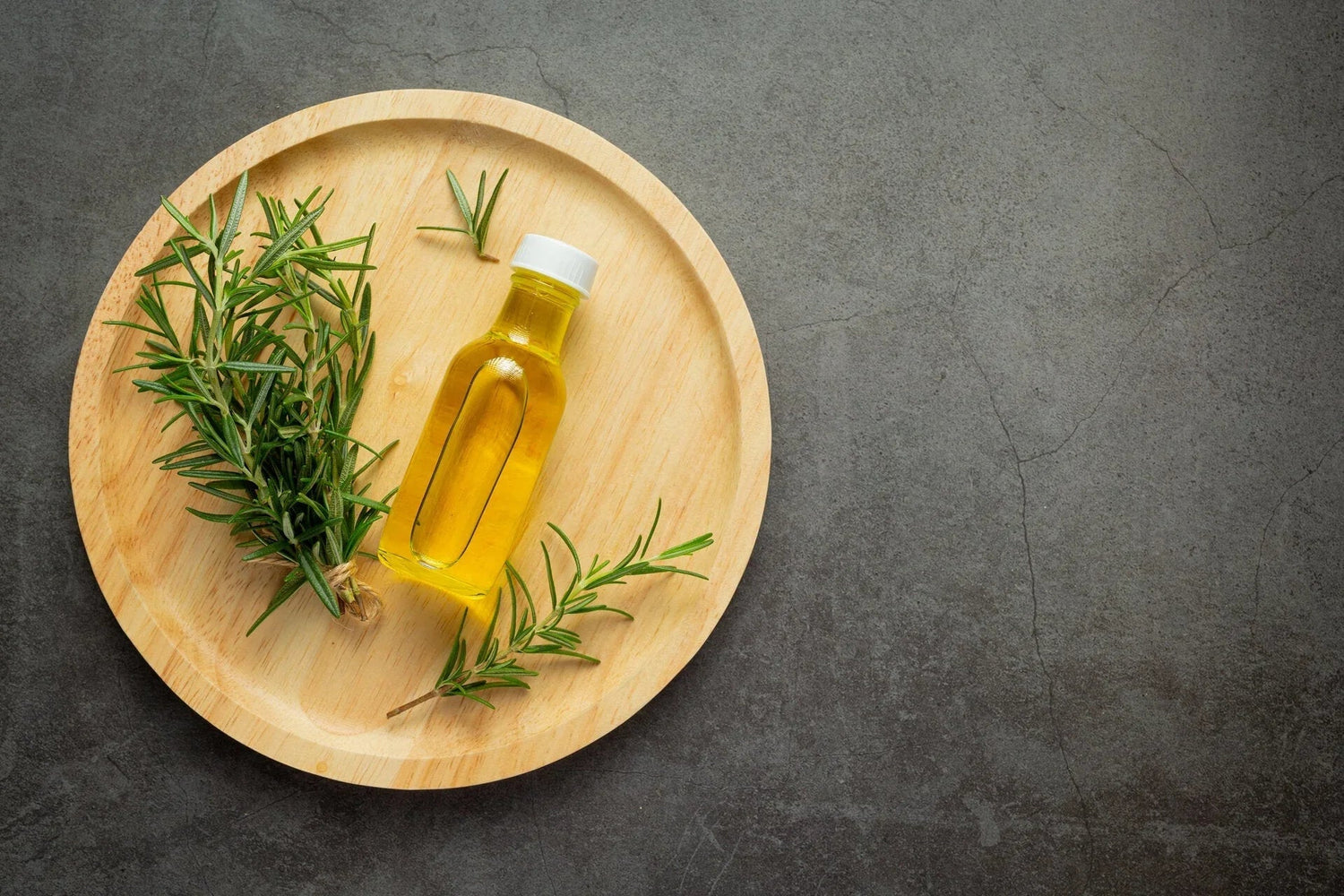 Small bottle of golden oil and fresh rosemary sprigs on a round wooden plate on concrete surface